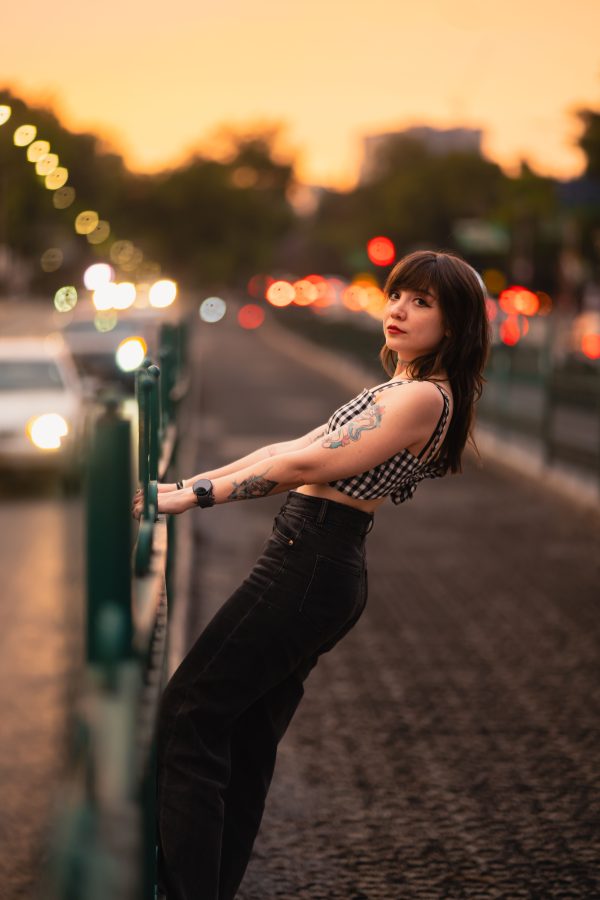 Stylish woman with tattoos leaning on city railing during a vibrant sunset. Mobile 4K