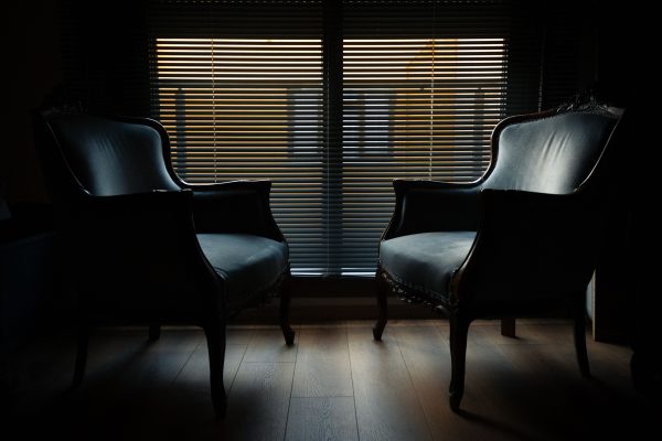Two vintage armchairs silhouetted against a window with jalousie blinds, creating a moody ambiance. Dark 4K