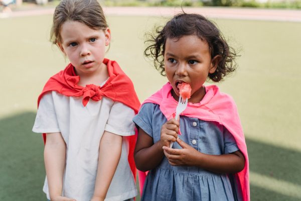 Two young girls wearing colorful capes, enjoying watermelon on a sunny day outdoors. Geek 4K