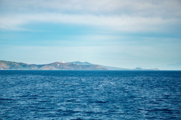 Vast ocean with distant island and lighthouse under a clear blue sky, offering a serene coastal landscape. Nature 4K