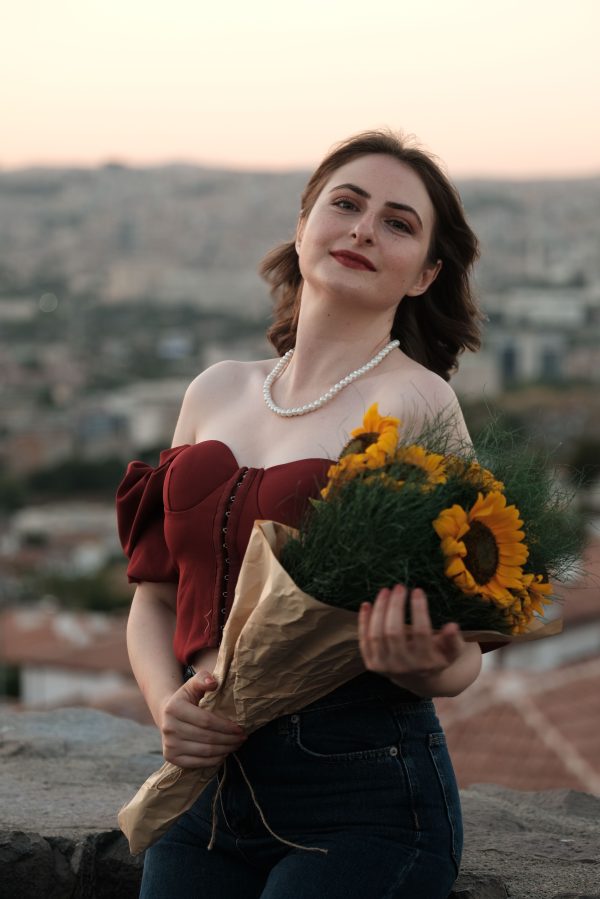 Woman in a stylish outfit holding sunflower bouquet against cityscape backdrop during sunset. Mobile 4K