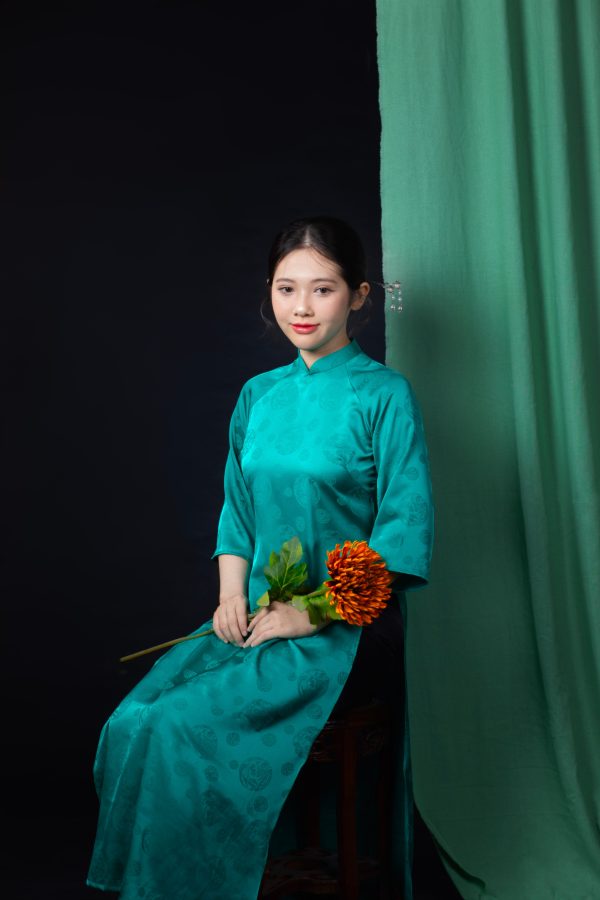 Young Vietnamese woman in a teal ao dai holding a flower, posing against a dark background. Mobile 4K