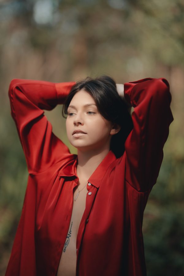 Young woman posing outdoors with hands behind her head wearing a red shirt. Mobile 4K